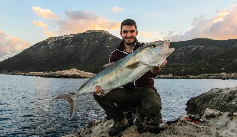 Angler crouching on the shore at dusk with a large silver fish, hills behind
